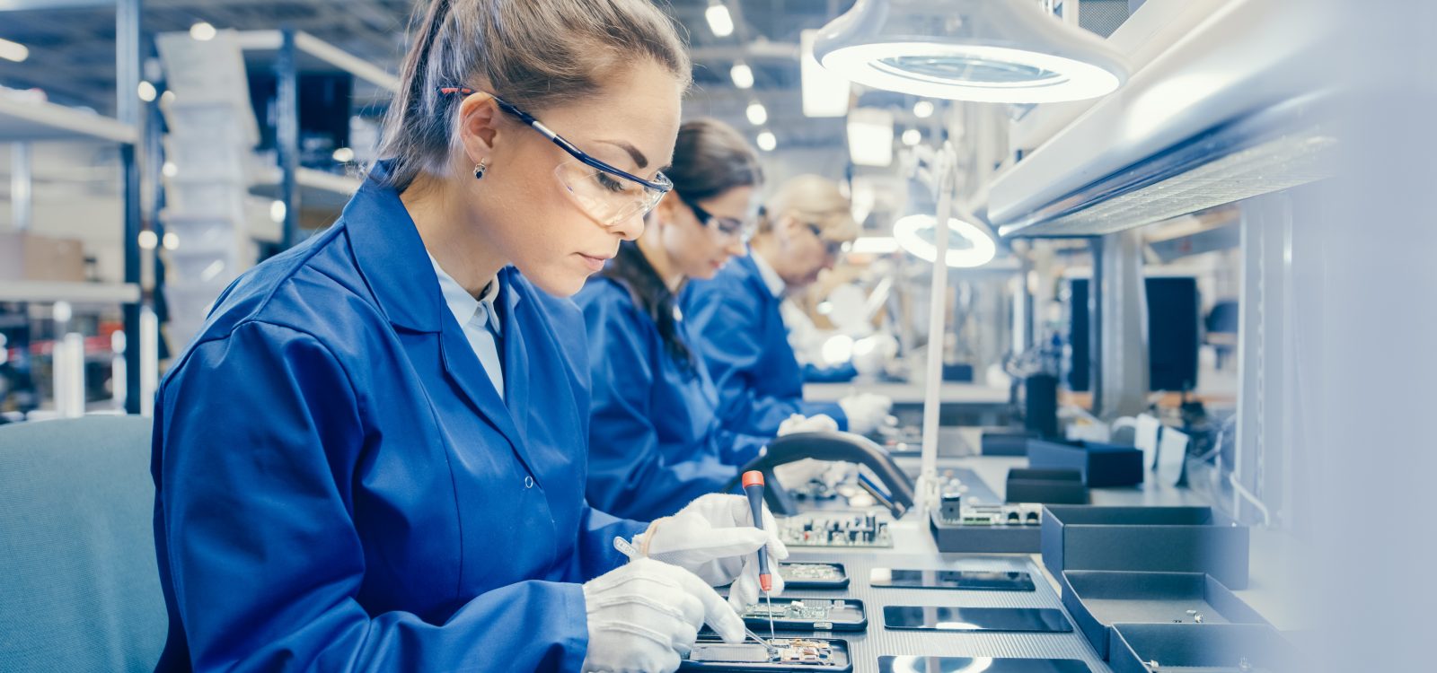 Woman Electronics Factory Worker in Blue Work Coat and Protective Glasses is Assembling Smartphones with Screwdriver. High Tech Factory Facility with more Employees in the Background.